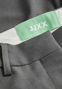 Pantalon en tissu gris avec une étiquette rectangulaire verte de la marque JJXX cousue à l'intérieur de la ceinture, avec des coutures blanches et une passants de ceinture.