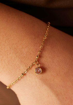 Gold bracelet chain with a small round purple gem pendant, resting on skin. Fine links show a delicate design and smooth texture.
