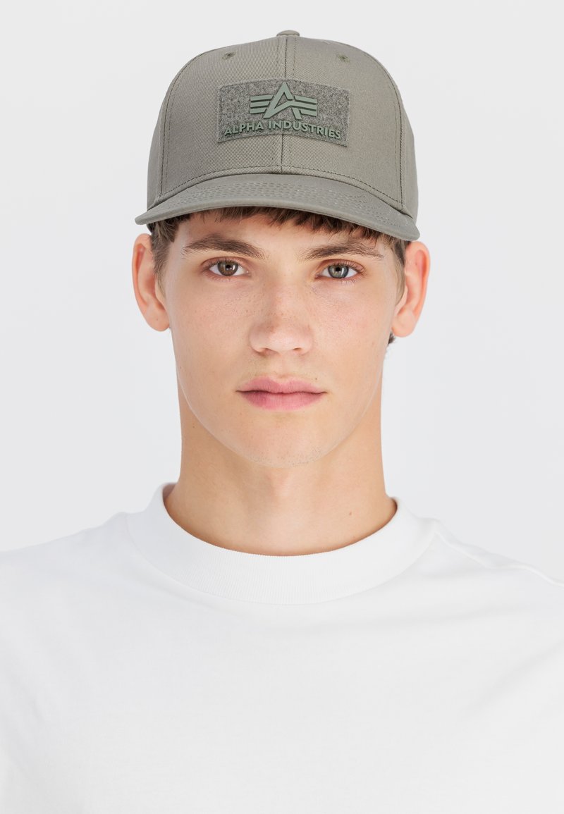 Junger Mann mit heller Haut, der eine graue Alpha Industries Kappe und ein weißes Rundhalsshirt trägt und mit neutralem Gesichtsausdruck nach vorne schaut.