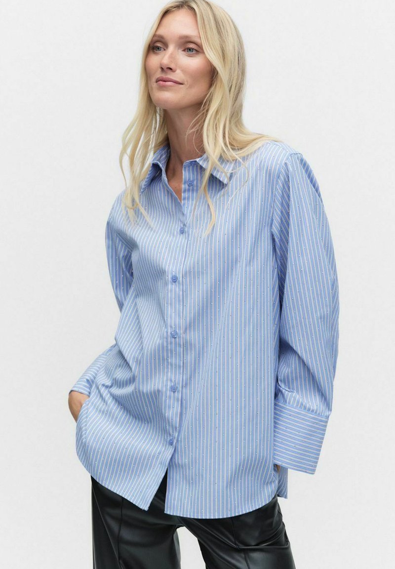 Ljusblå skjorta med knappar, vertikala vita ränder, långa ärmar och en avslappnad passform. Har en krage och två framfickor.