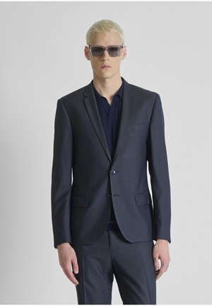 Jonge man met kort blond haar, donkerzonnebril, een marineblauwe blazer en bijpassende broek, tegen een effen lichte achtergrond.