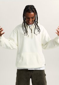 Felpa bianca con cappuccio e tasca frontale, con patch del logo tonal; vestibilità ampia con maniche lunghe, indossata sopra una maglietta grigia e pantaloni scuri.