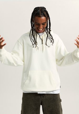 Sweat-shirt à capuche blanche avec une poche avant et un patch de logo de la marque ton sur ton ; coupe ample avec des manches longues, porté sur une chemise grise et un pantalon sombre.