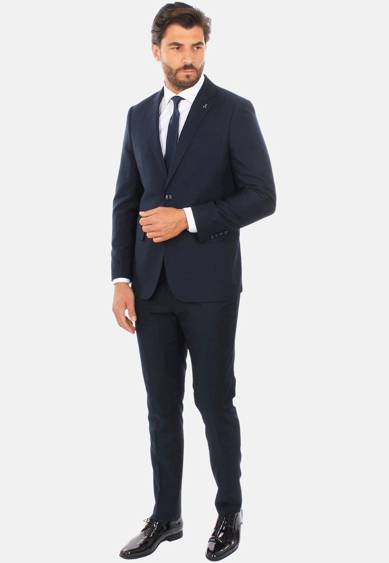 Completo blu con un solo bottone, design slim fit, tessuto strutturato, abbinato a una camicia bianca e una cravatta blu, completato da scarpe nere lucide.