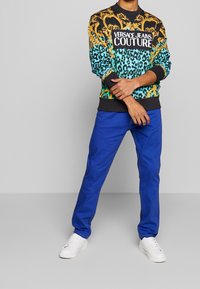Sudadera negra y turquesa con estampado y logo, combinada con pantalones azules y zapatillas blancas. Presenta un diseño gráfico y colores contrastantes.