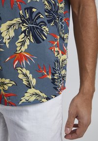 Hawaïaans shirt met een blauwe achtergrond en groene, gele en rode bladranden. Gekleed met witte linnen shorts, die een lichte textuur tonen.