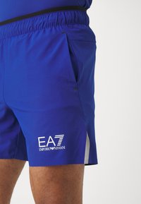 Modré atletické šortky z ľahkého materiálu. Obsahujú elastický pás, bočné vrecká a logo EA7 v bielej farbe. Doplnkové strieborné detaily.