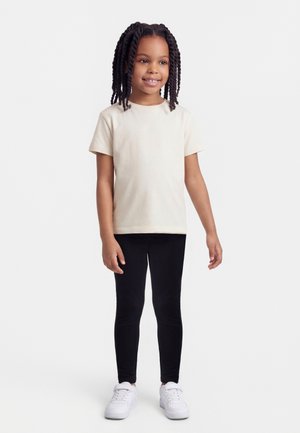 Niña con cabello trenzado que lleva una camiseta beige de manga corta, leggings negros y zapatillas blancas, de pie y sonriendo sobre un fondo blanco.