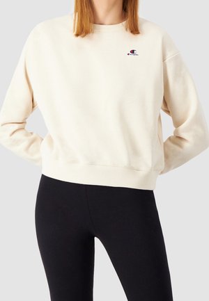 Personne portant un sweat-shirt crème Champion avec un petit logo sur la poitrine, associé à des leggings noirs, les mains posées sur les hanches, sur un fond uni.