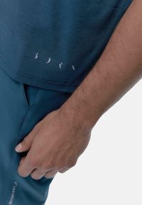 Ciemnoniebieska koszula krótki rękaw o gładkiej fakturze z białym nadrukiem na dole. Powyżej znajduje się ręka oparta na turkusowych spodniach.