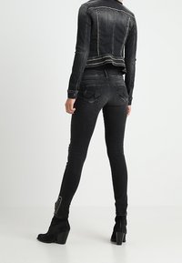 Osoba v přiléhavé černé džínové bundě s hroty, ladících skinny džínech a černých kotníkových botách na podpatku, stojící na jednobarevném pozadí.