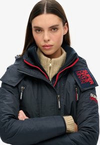 Veste matelassée marine avec des accents rouges, deux poches zippées et des écussons de marque bien visibles. Portée par-dessus un pull à col roulé beige avec une texture côtelée.