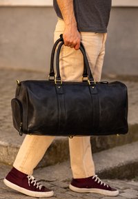 Sort læder duffel bag med guldfarvet hardware, rund form, glat overflade, top-håndtag og sidelomme. En person holder den, mens de går.