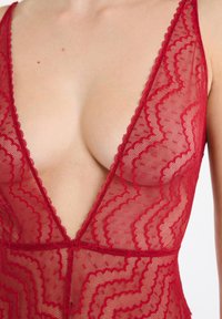 Body en dentelle rouge avec un décolleté plongeant en V, bordure festonnée et motif en zigzag. Un tissu délicat et transparent met en valeur son design texturé.