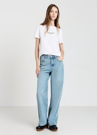 T-shirt en coton blanc avec un design brodé, associé à un jean large bleu clair et des chaussures à plateforme noires. Tenue simple et décontractée.