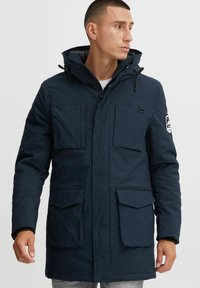Marineblauer Parka mit Kapuze, mehreren vorderen Taschen, Reißverschluss und kontrastierender grauer Innenauskleidung. Verfügt über verstellbare Kordelzüge.