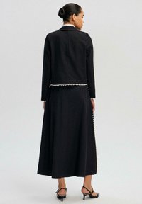 Černý dvoudílný outfit složený z krátké bundy s vlnitým okrajem a dlouhé sukně s bočními detaily. Hladká tkanina a přiléhavý střih.