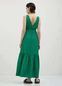 Grün ärmelloses Kleid mit V-Rücken, elastischem Taillenbund, geschichteter Rock und strukturiertem Stoff, der bis zu den Knöcheln reicht. Getragen mit flachen schwarzen Schuhen.