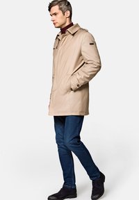 Beżowy wodoodporny płaszcz o klasycznym designie, z dużym kołnierzem, zapinanymi mankietami i bocznymi kieszeniami, noszony na ciemnym swetrze i niebieskich dżinsach.