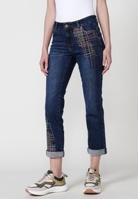 Modri jeans s zvitim robom, okrašeni z večbarvnimi vezeninami na levem stegnu. Ustrezno kombinirani s športnimi supergami.