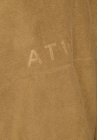 Brun mocka med en mjuk textur. Har subtila sömmar och präglad "ATV"-logotyp, vilket visar en minimalistisk design.