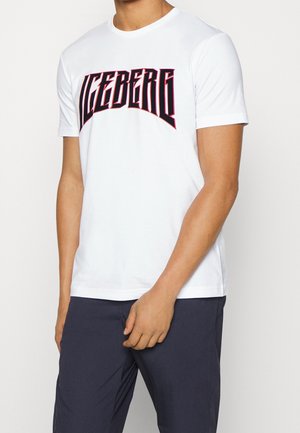 Homme portant un t-shirt blanc à manches courtes avec "ICEBERG" imprimé en lettres stylisées et en gras sur la poitrine, ainsi qu'un pantalon sombre.
