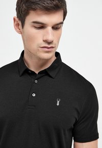 Schwarzes Polohemd mit Kragen, zwei Knöpfen und einem kleinen grauen Hummeremblem auf der linken Brust. Weicher Stoff mit glatter Textur.