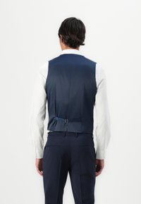 Marineblaues ärmelloses Oberteil mit verstellbarem Rückengurt, getragen über einem weißen Hemd. Glattes Material, minimales Design, mit sichtbaren dunklen Hosen.