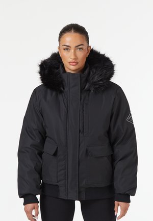 Mujer con chaqueta de invierno negra con capucha forrada de piel, de pie mirando al frente sobre un fondo blanco liso.