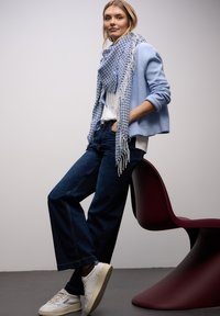 Jasnoniebieski kardigan na białym topie, niebiesko-biała wzorzysta apaszka, ciemne dżinsy o szerokich nogawkach oraz białe trampki. W tle bordowy fotel.