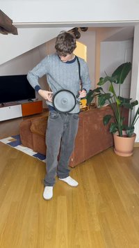 Altavoz circular negro con un asa, colocado a la altura de la cintura. El usuario lleva un suéter gris con un patrón y jeans oscuros. Entorno interior con plantas.