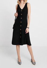 Schwarzes geripptes Kleid mit V-Ausschnitt, goldenen Knöpfen entlang der Vorderseite und einem fließenden Rock. Getragen mit gemusterten High Heels.