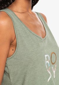 Vihreä tanktoppi, jossa on V-niskasuunnittelu, valmistettu pehmeästä materiaalista, ja jossa on kukkakoristeinen "ROXY"-logo erottuvissa väreissä.