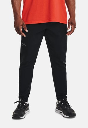 Homme portant un pantalon de sport noir et des chaussures de course noires, debout les pieds légèrement écartés sur fond blanc.