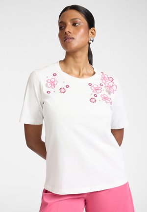 Vrouw draagt witte top met korte mouwen met roze bloemrijk borduurwerk op de schouders en roze broek, staand tegen een effen witte achtergrond.