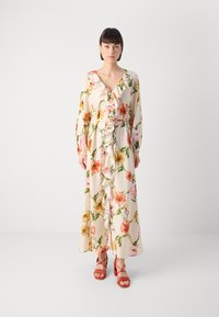 Abito maxi floreale con base crema, maniche lunghe, davanti arricciato e vita con cintura. Abbinato a sandali rossi con cinturini.