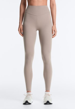 Woman wearing high-waisted beige leggings and white athletic shoes, standing against a plain light background.