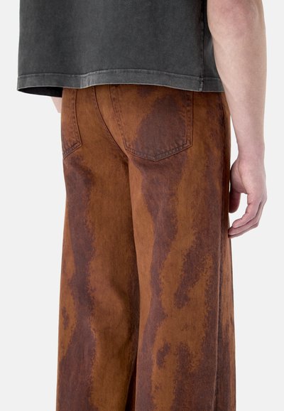 Mann trägt braune, weit geschnittene Hose mit dunklen, wellenförmigen Mustern und ein graues T-Shirt, dargestellt von der Taille abwärts vor weißem Hintergrund.