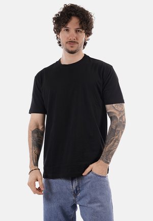 Uomo con capelli ricci che indossa una t-shirt nera e jeans blu, che mostra tatuaggi su entrambi gli avambracci, in piedi davanti a uno sfondo semplice.