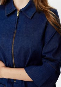 Tamsiai mėlyna džinsinė striukė su užtrauktuku priekyje, klasikinio kirpimo apykakle ir trijų ketvirčių rankovėmis su kontrastingu siuvimu.