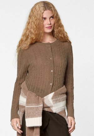 Vrouw met krullend blond haar, gekleed in een bruin gebreid vest met knopen en een beige-wit gestreepte gebreide sjaal gestrikt om haar middel.