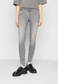 Pantalones vaqueros ajustados grises con detalles desgastados en las rodillas, combinados con zapatos negros gruesos. El material parece vaquero con una textura suave.