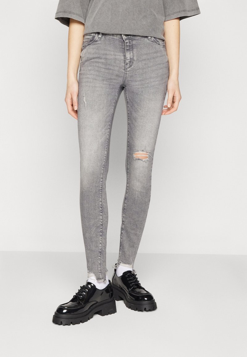 Pantalones vaqueros ajustados grises con detalles desgastados en las rodillas, combinados con zapatos negros gruesos. El material parece vaquero con una textura suave.
