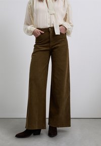 Pantalon large à taille haute en velours côtelé marron à la texture douce. Stylisé avec un chemisier léger et des bottes à talons foncées, vue de face.