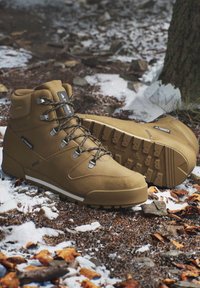 Brown high-top hiking boots with rugged soles resting on snowy, rocky forest ground near a tree trunk.