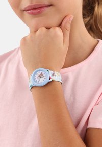 Blauwe horloge met een stofband met bloemenpatroon, voorzien van een ronde witte wijzerplaat en paarse uurmarkers, gedragen om een pols. Roze shirt op de achtergrond.