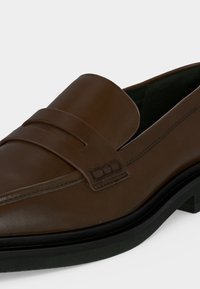 Mocassin en cuir marron avec une large bride et des détails cousus sur la partie supérieure, avec une semelle en caoutchouc noire et un petit talon.