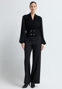 Sort, langærmet top med indpakningsdesign og guldknapdetaljer. Sat sammen med skræddersyede, vide bukser og sorte spidse hæle.