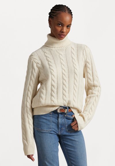 Chunky cream kabelbrei trui met een hoge, omgeslagen kraag; geribde manchetten en zoom; gedragen met blauwe denim jeans en een bruine riem.