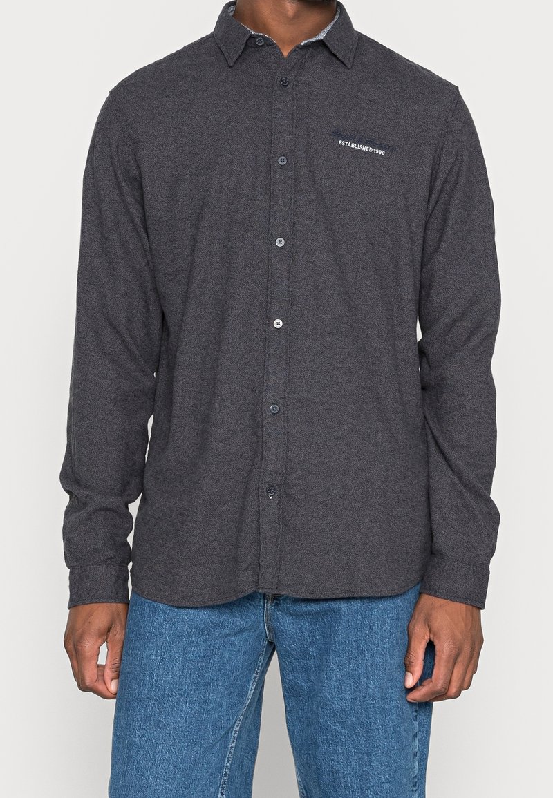 Węgielnoszara koszula zapinana na guziki, wykonana z tkaniny o fakturze, z długimi rękawami i granatowym kołnierzem. Na piersi znajduje się małe haftowane logo.